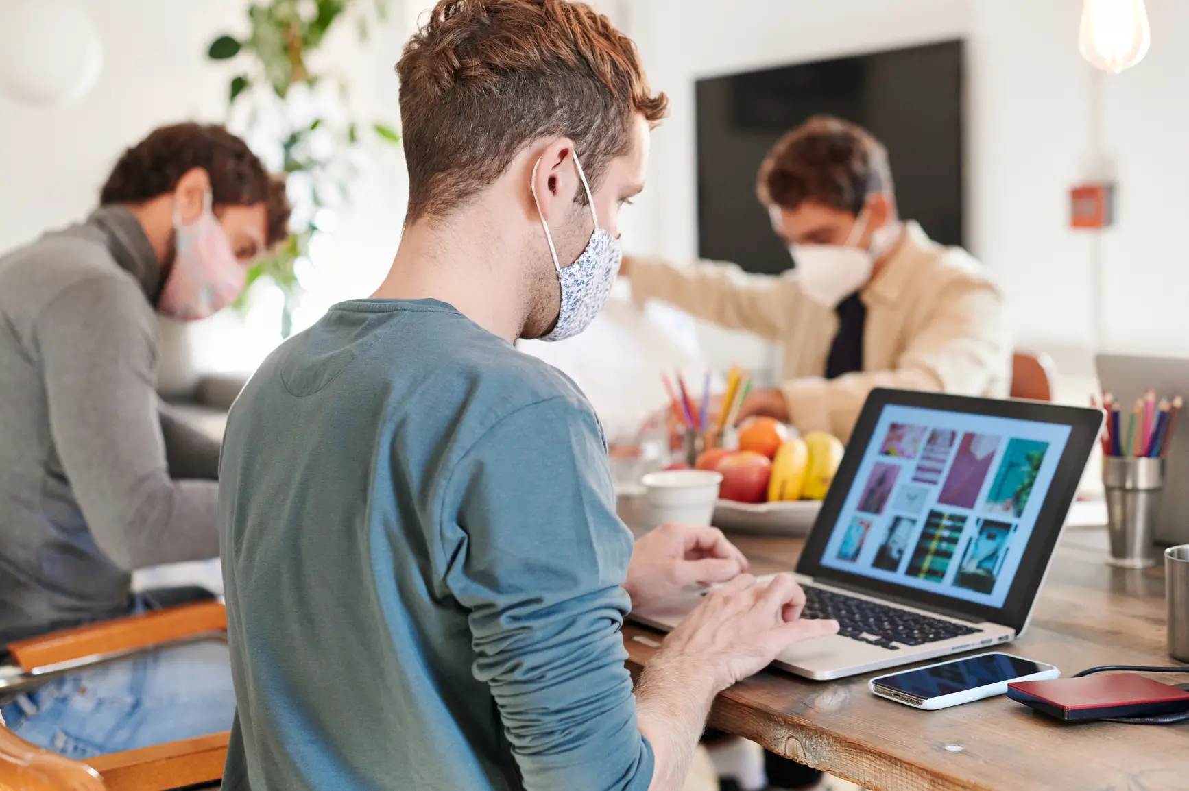 Ein junger Designer arbeitet am Laptop, er und seine Kollegen im Hintergrund tragen Gesichtsmasken.