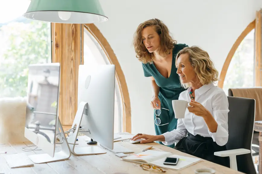 A woman is standing behind her colleague, who is sitting at her desk. Together they look into the stand PC.