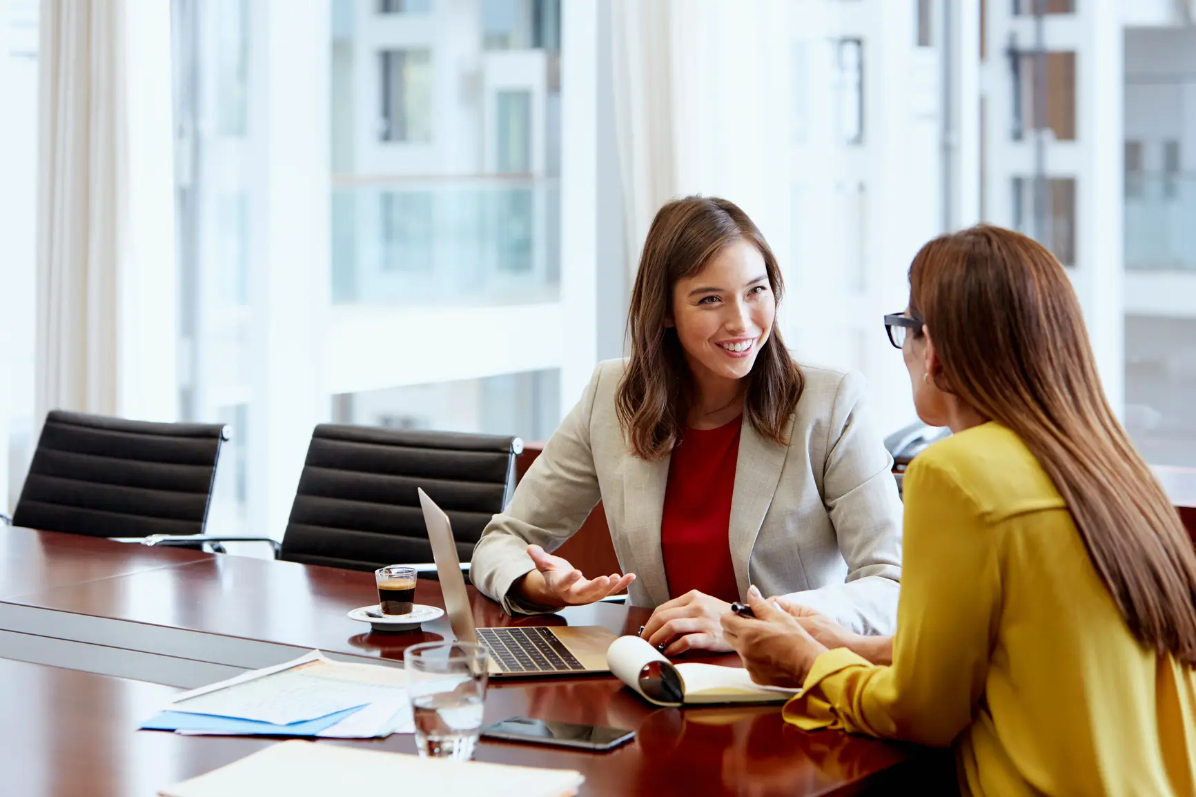 Two women - one is seen frontally, one from behind - are talking to each other at a conference table.