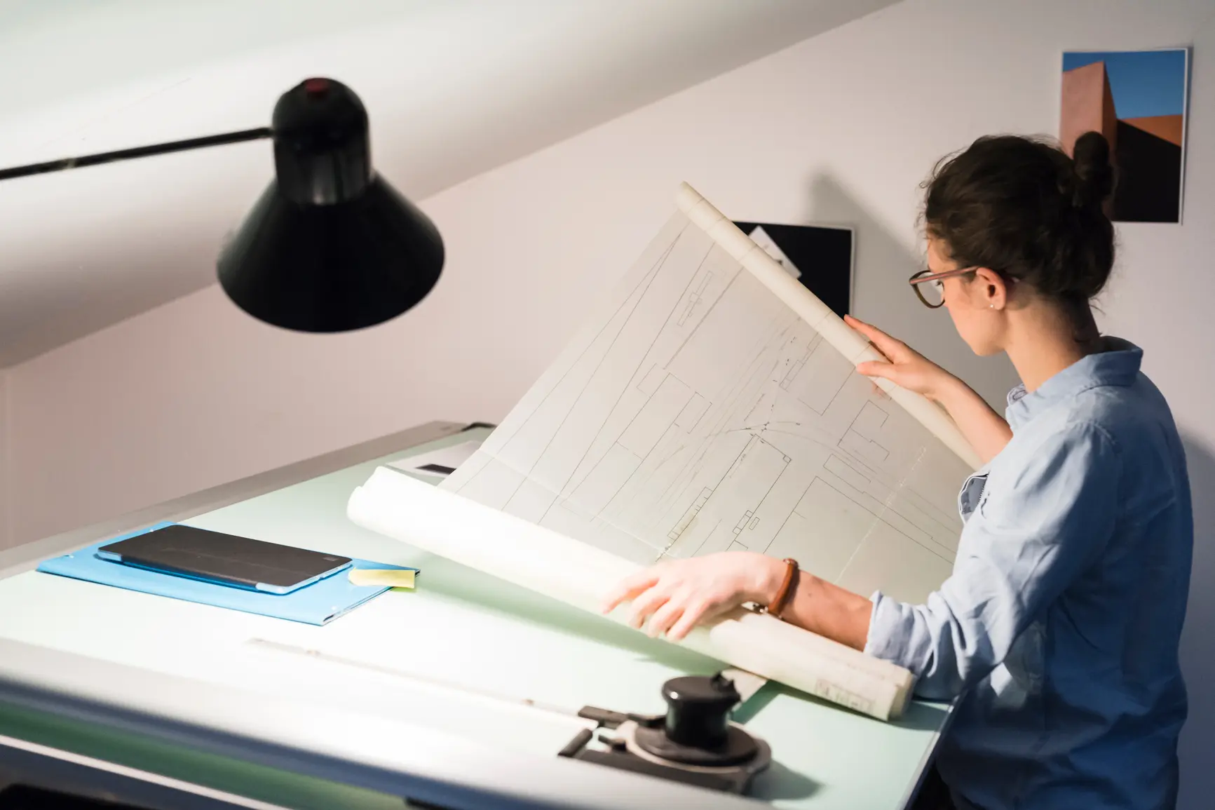 An architect sits at her desk and unrolls a large blueprint in front of her.