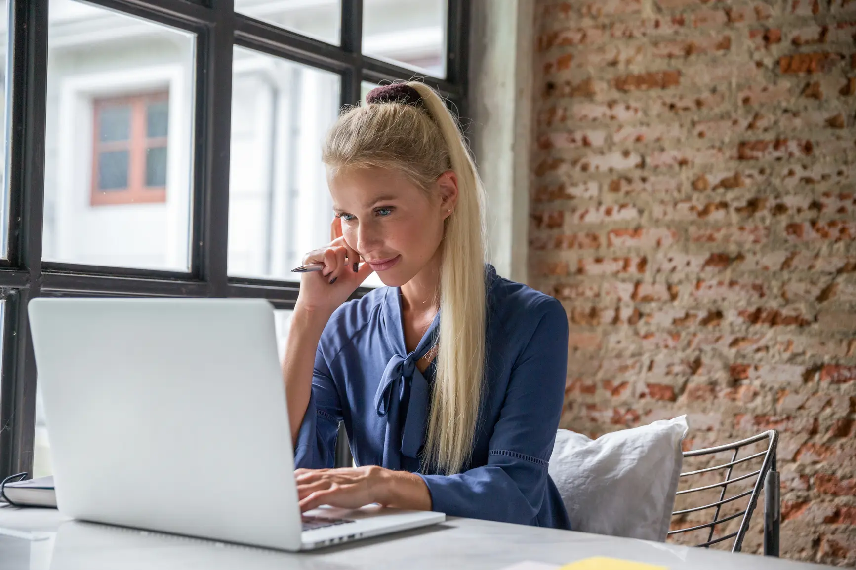 A young woman sits in front of her laptop with one hand on her laptop, one hand on her face. In the background you can see a wall.