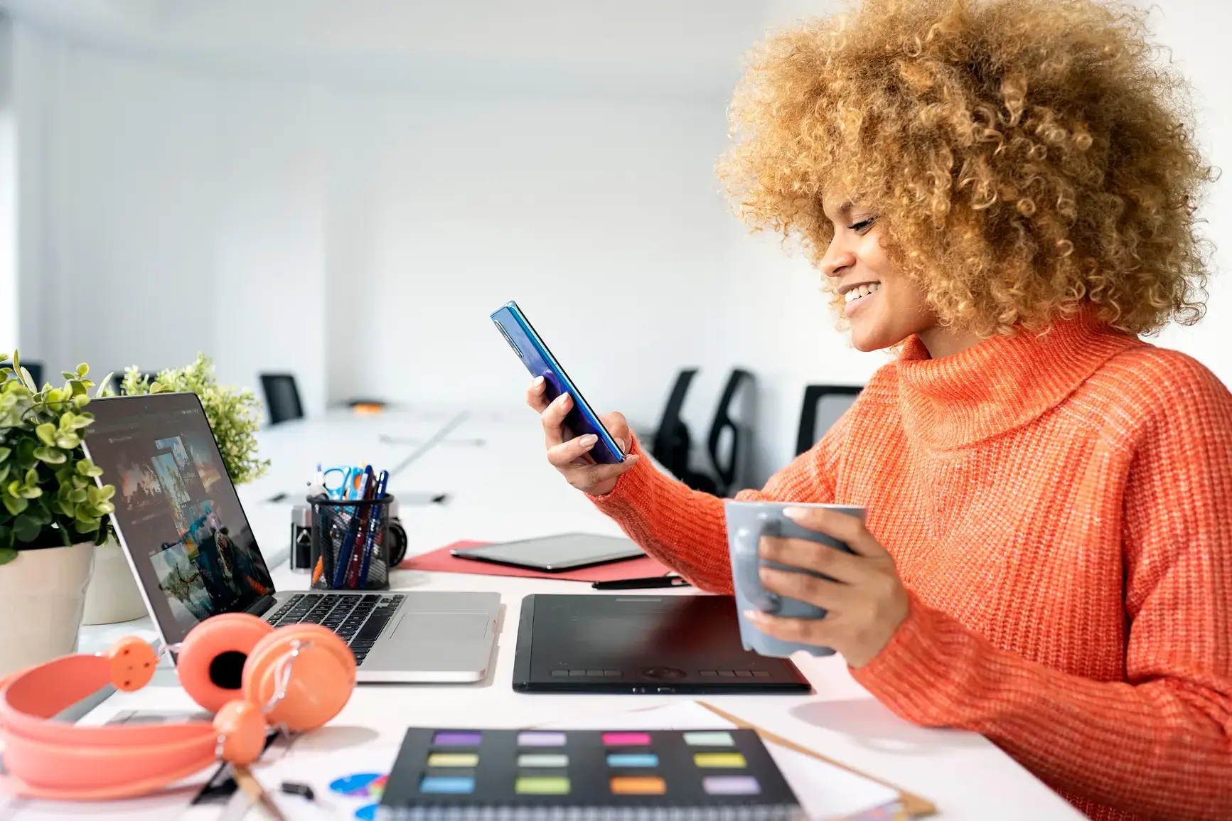 A woman sits at her desk with coffee in hand in front of her laptop and drawing board. In her hand she holds a smartphone.