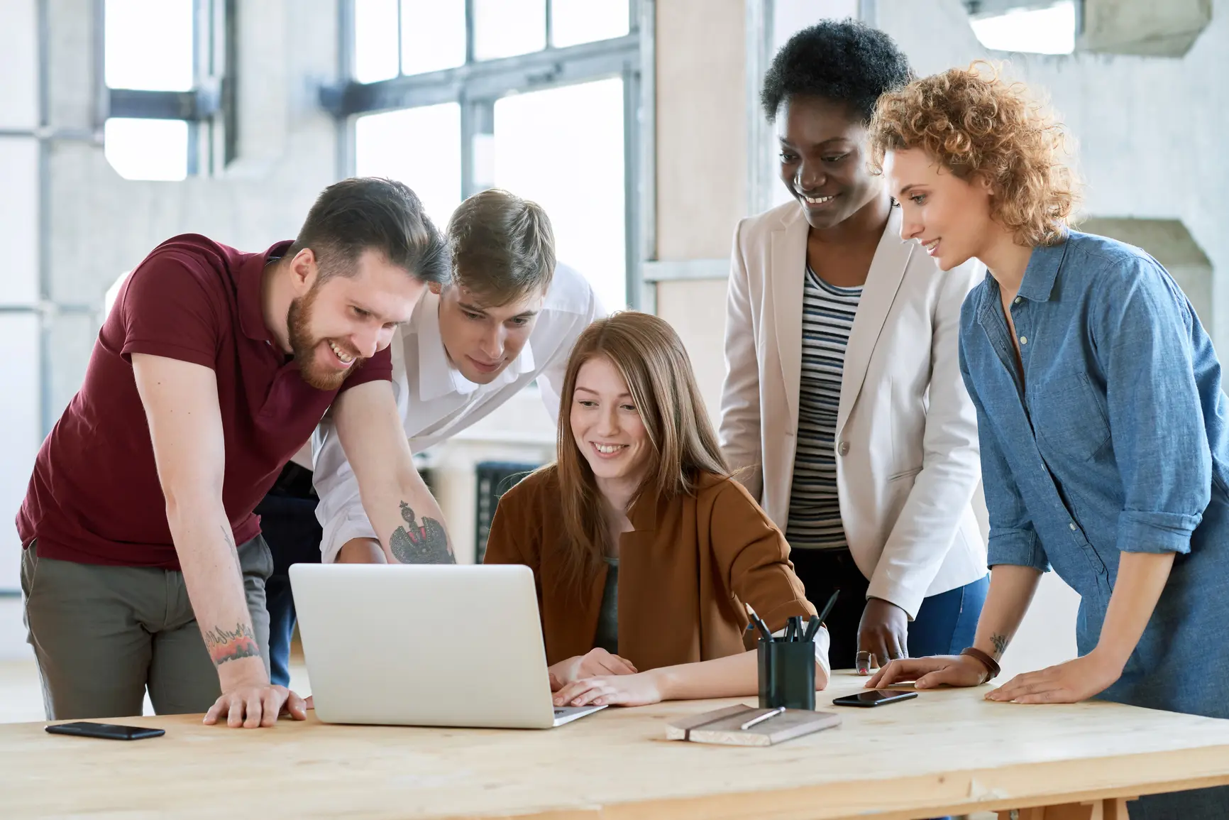 A woman is sitting at her desk in front of her laptop. Around her, four colleagues have gathered to watch something together.