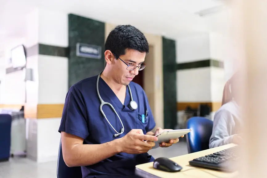 A doctor with a stethoscope around his neck stands in front of a tablet in a hospital.