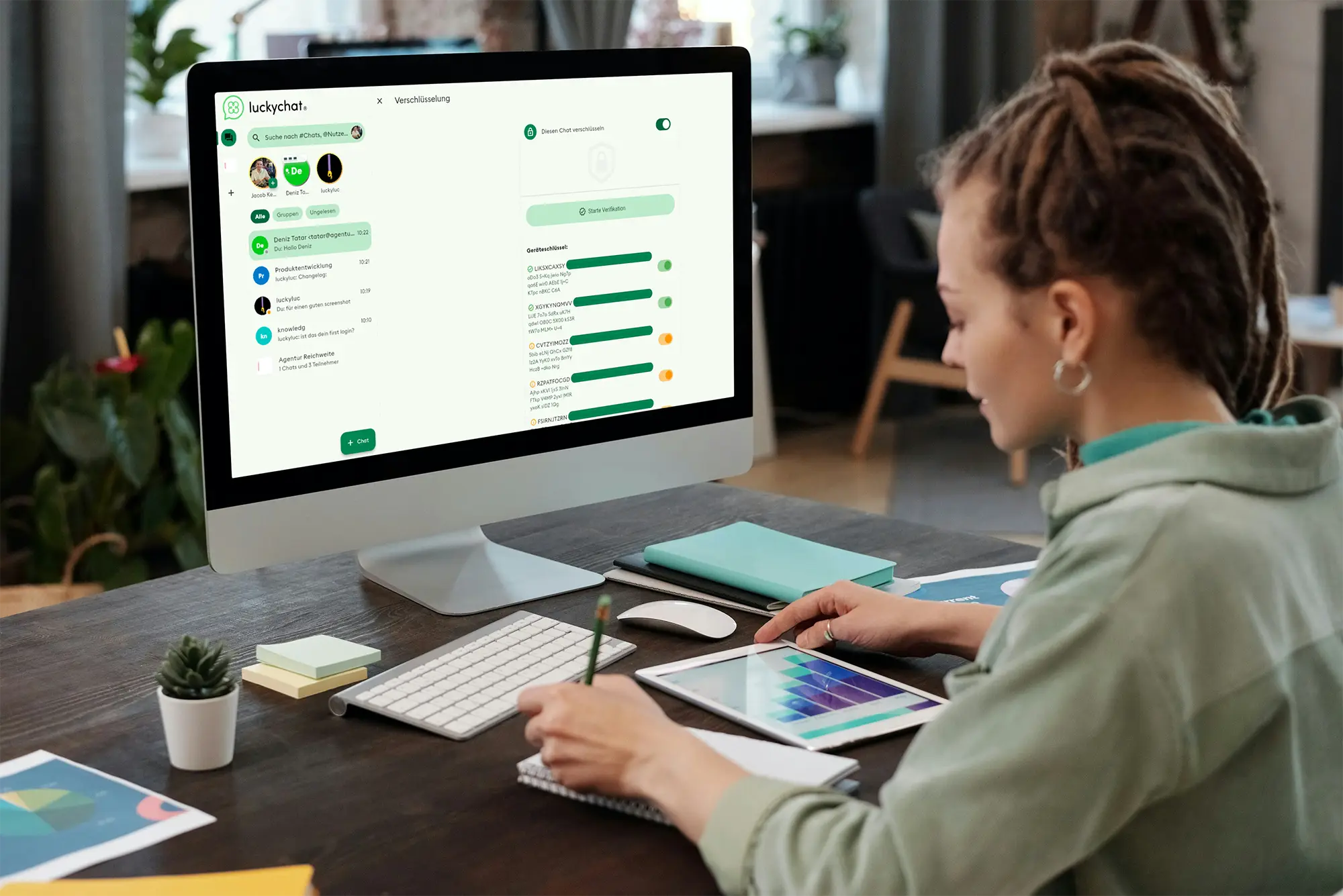 Woman working at a desk with a desktop computer displaying the luckychat messaging application. She is simultaneously using a tablet for graphic design.