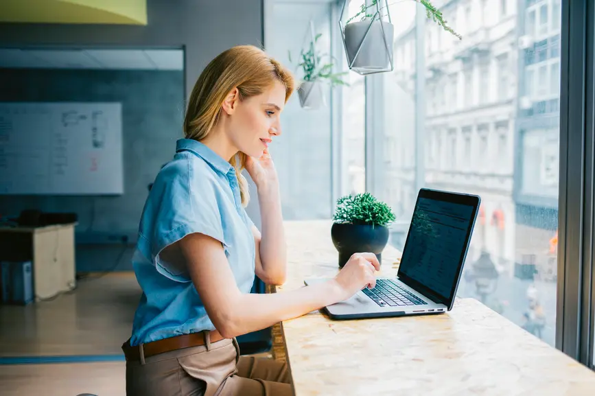 Eine Frau arbeitet am Laptop vor einer hellen Fensterfront.