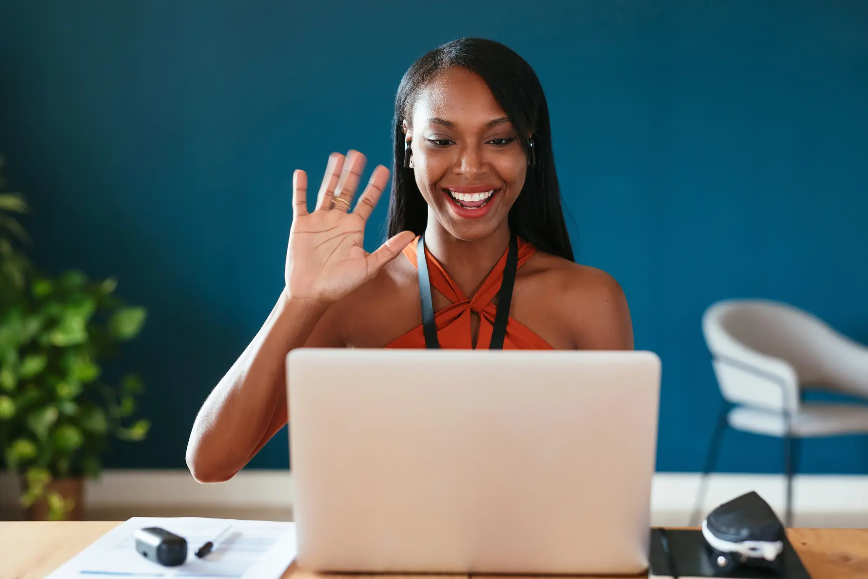 A woman sits frontally in front of her laptop. She waves and laughs into the camera.