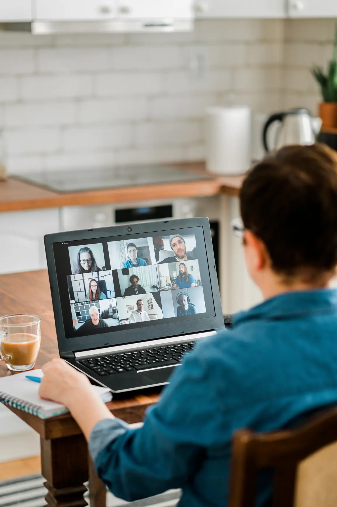 View over the shoulders of a woman working from the home office. On the table in front of her is a laptop with pictures of colleagues (video conference).