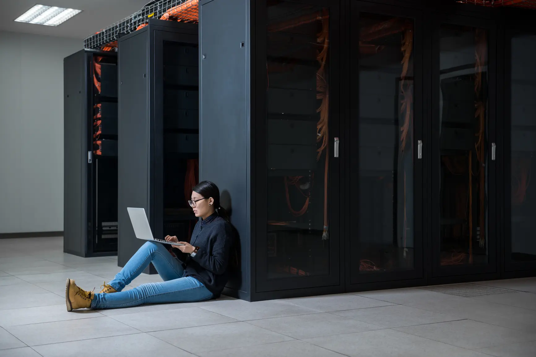 A woman sits with her laptop on the floor of a server room.