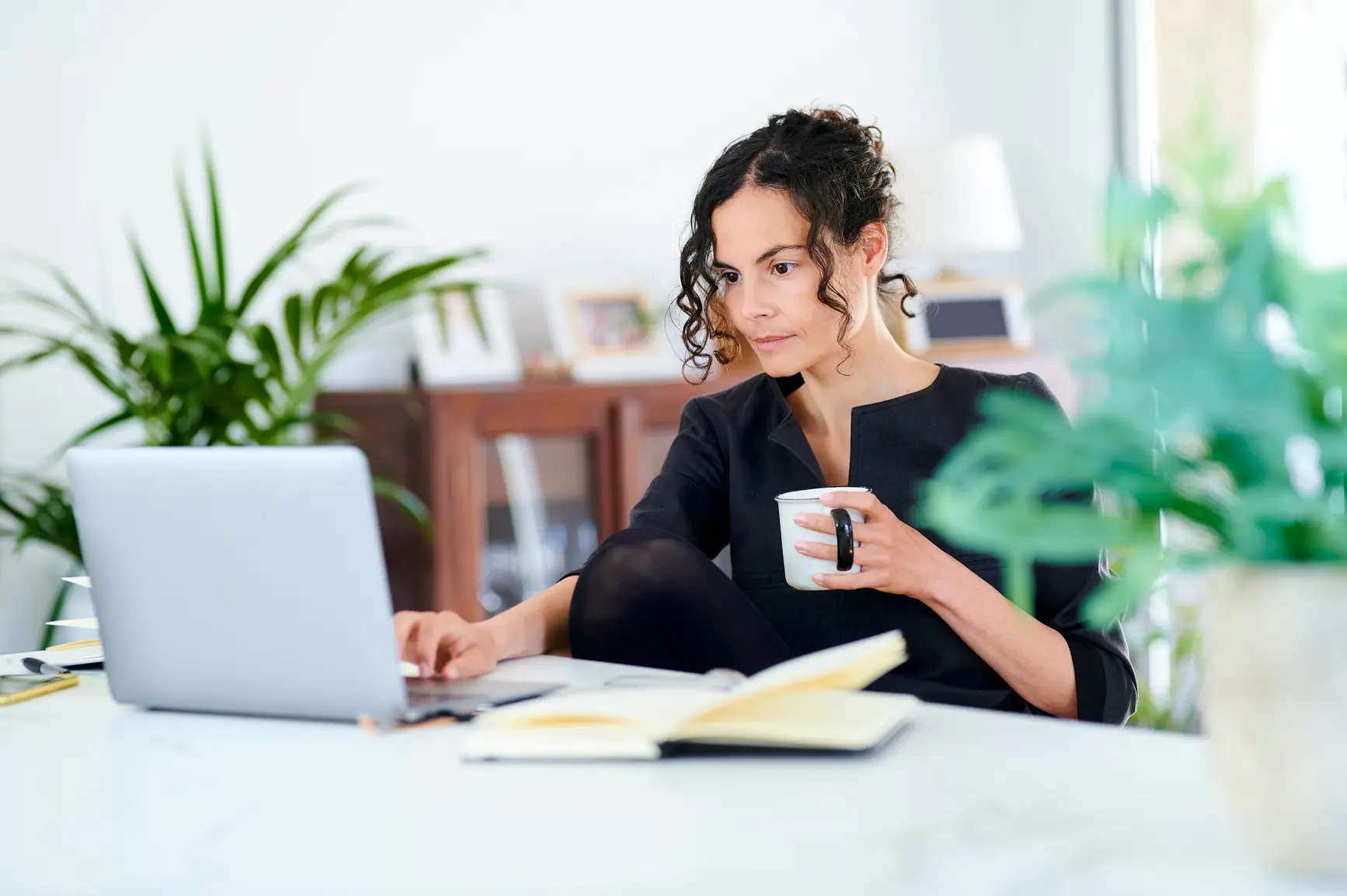 Eine Frau sitzt mit Kaffeetasse in der Hand vor ihrem Laptop. Daneben liegt ein Notizbuch.