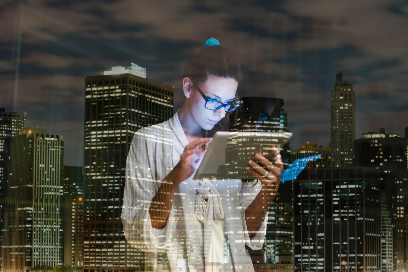 A woman looks at her tablet at night, the skyline of a city can be seen in the background.