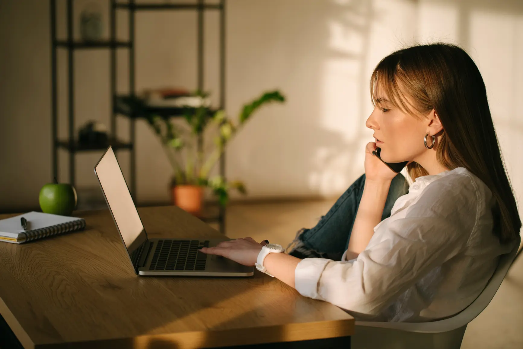 Eine Frau sitzt bei angenehmen Morgenlicht am Laptop und telefoniert.