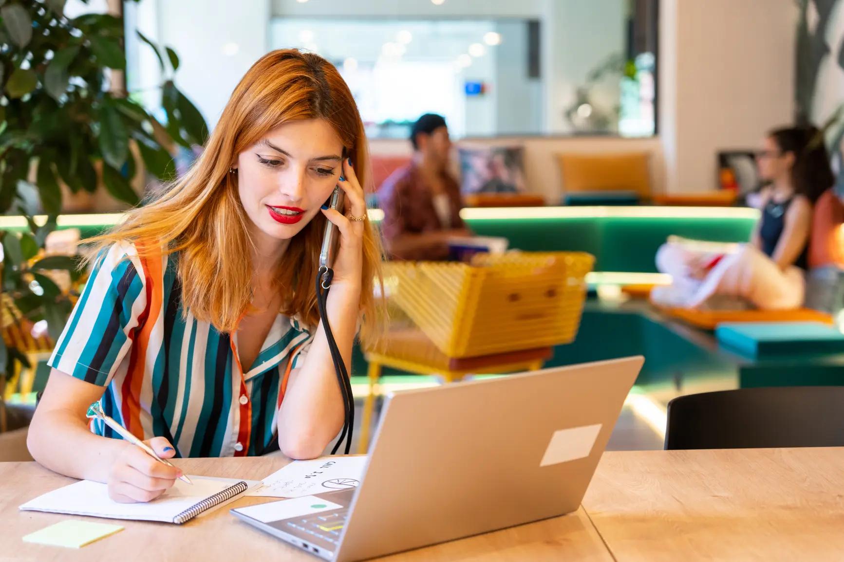 A woman is talking on the phone in front of her laptop and taking notes at the same time.