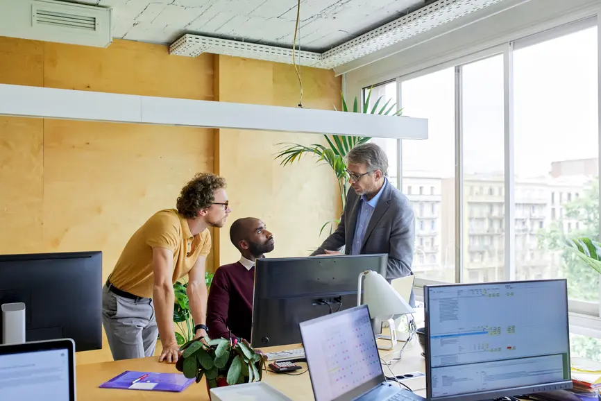 Three businessmen confer at a desk in the bright office.