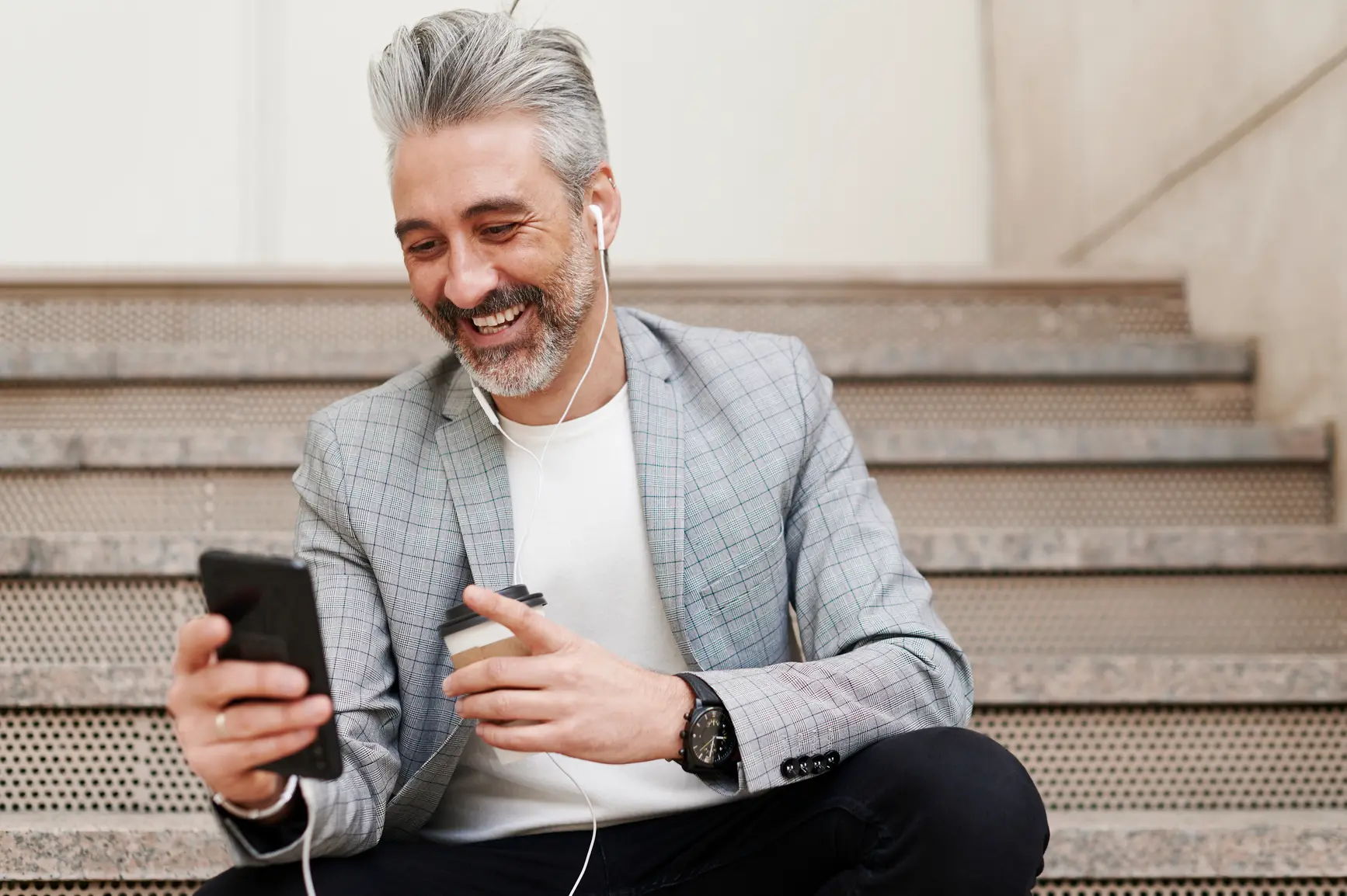 A businessman sits on a staircase talking on the phone with headphones in his ear and smartphone in his hand.