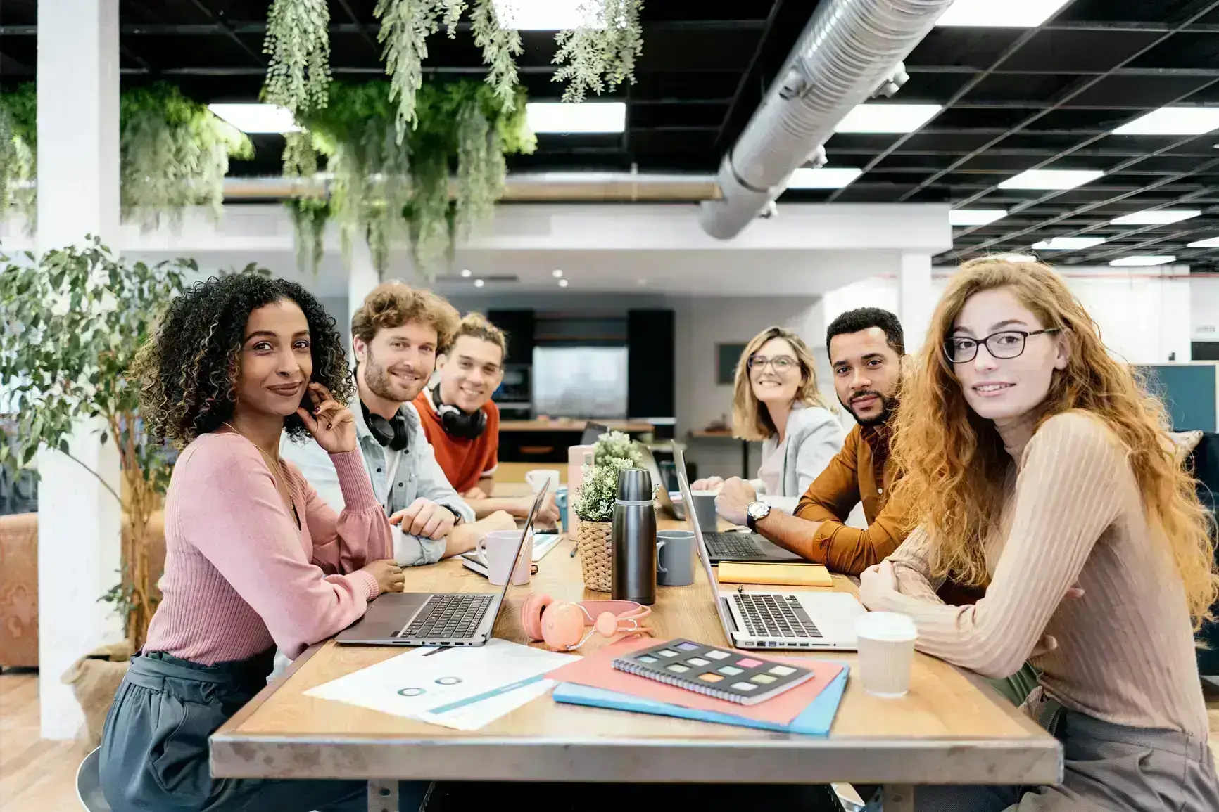 A team of six colleagues sits at a table in front of them laptops, drinks and notebooks. All of them are looking toward the camera.