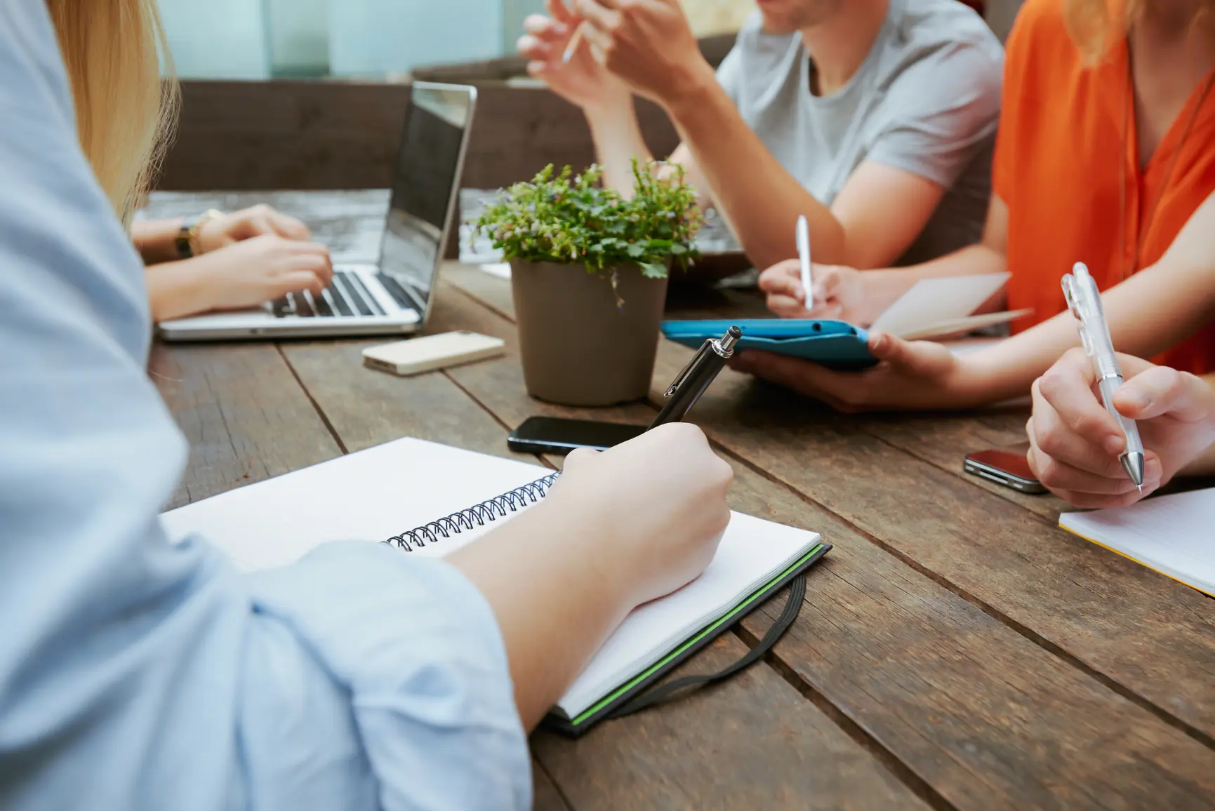 Junge Unternehmer befinden sich in einem Gruppenmeeting und machen Notizen. Zu sehen ist der Ausschnitt des Tisches mit den Haenden und der einzelnen Personen.