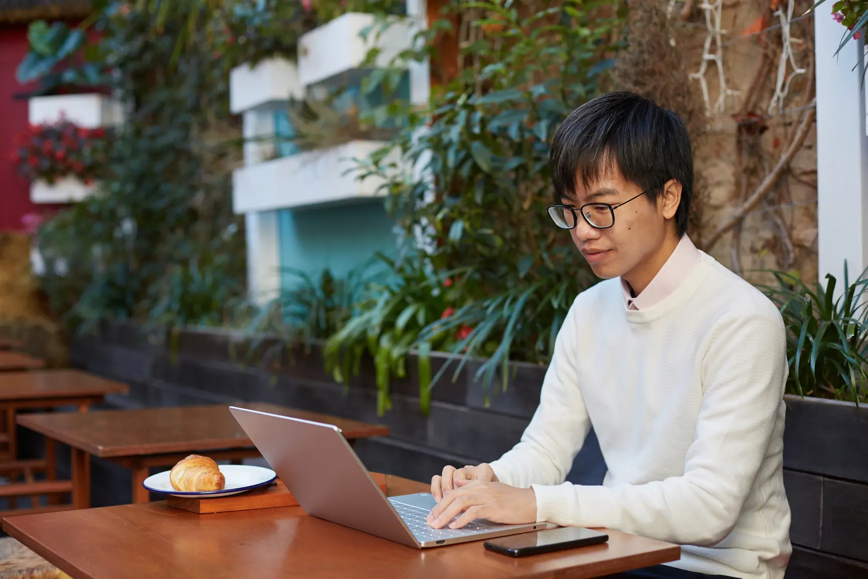 A young man is sitting at a table in the open. In front of him is his laptop and a plate with a croissant.