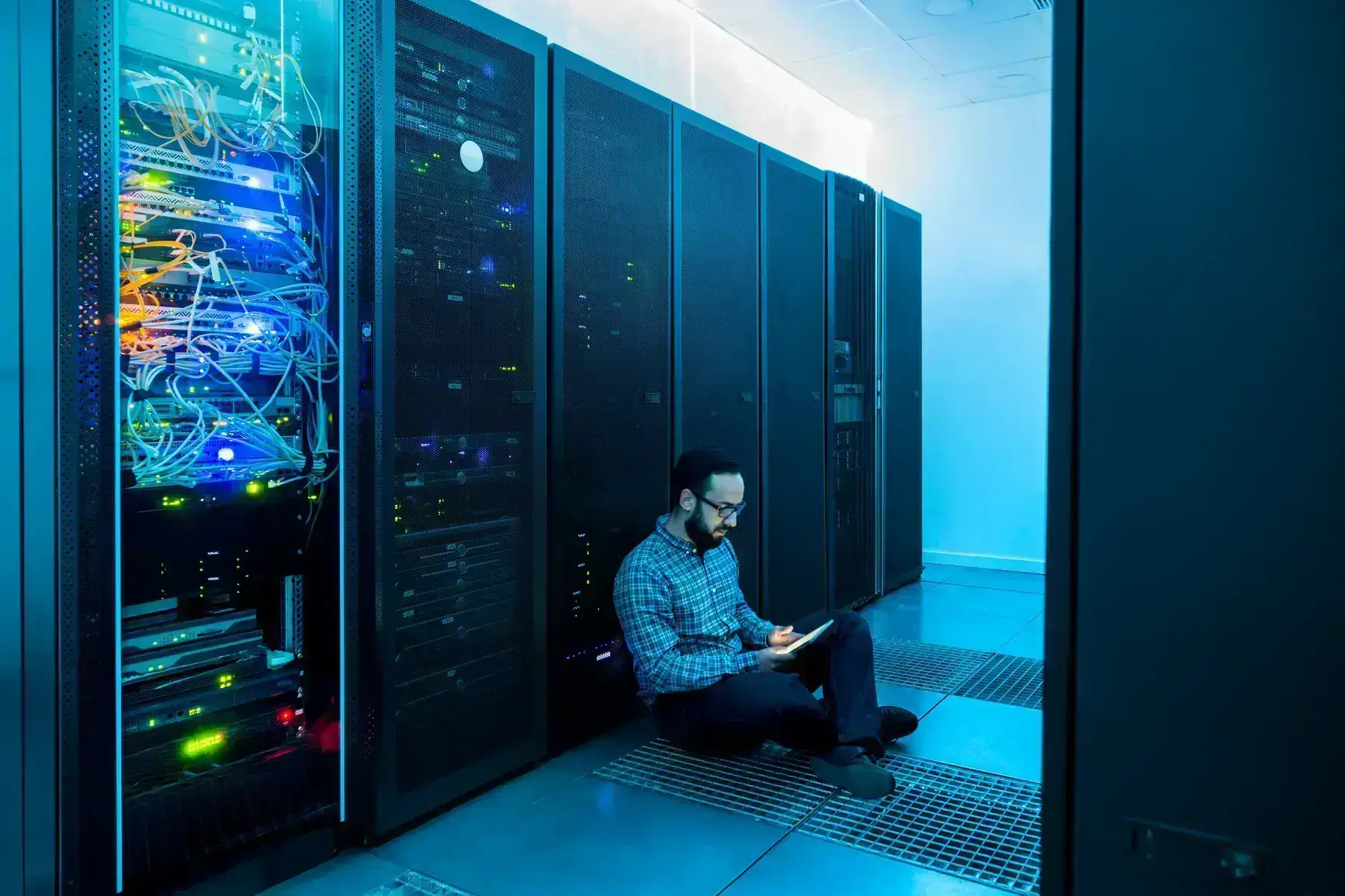 A man sits on the floor of a server room with his tablet.