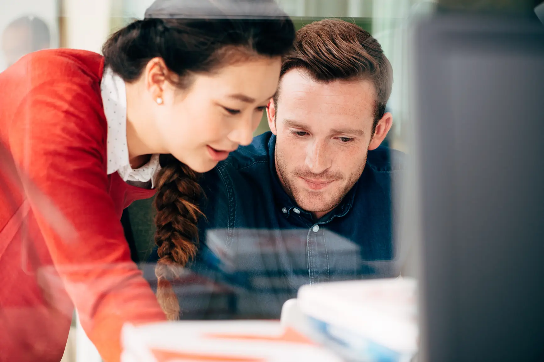 A woman leans down to a man sitting at his desk. Together they look at a document.