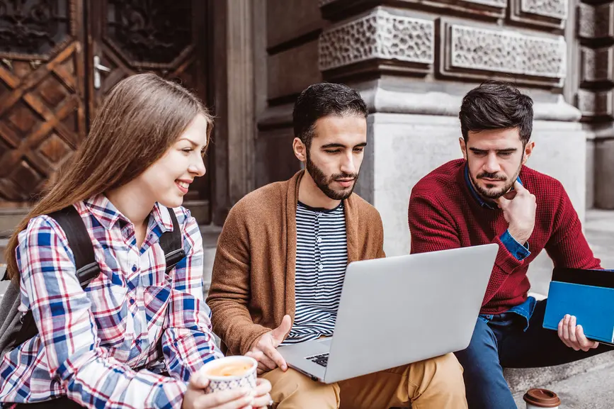 Three students are sitting outside on a staircase, working together on a laptop.