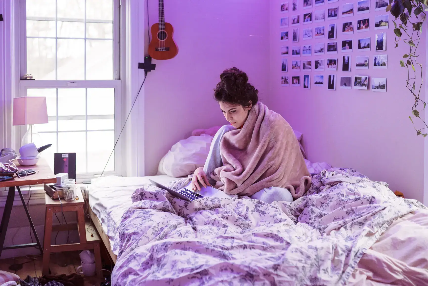 A student sits on her bed, snuggled in blankets, studying on her laptop.