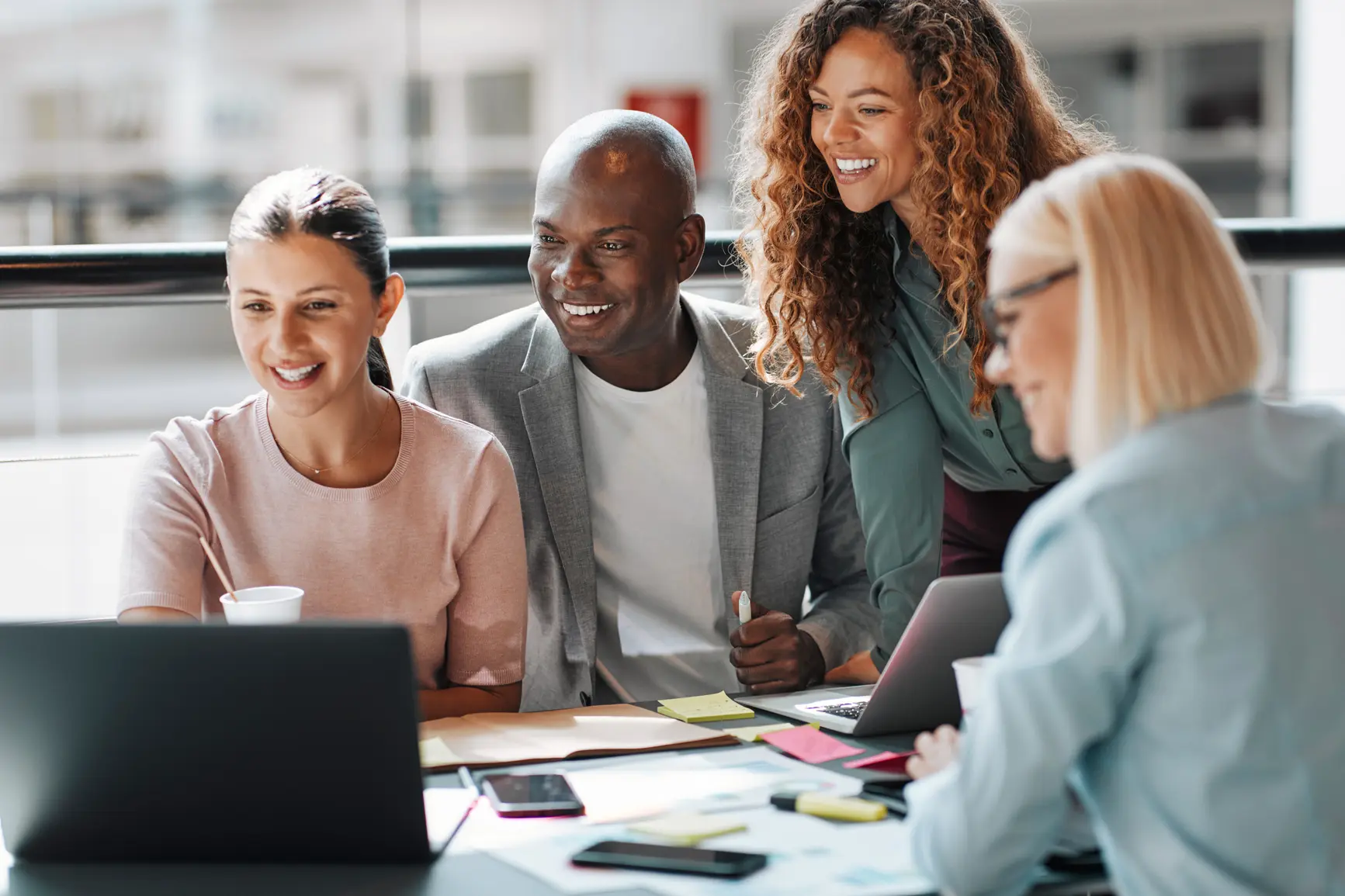 A team of four people gazes joyfully together into a laptop on a crowded work table.