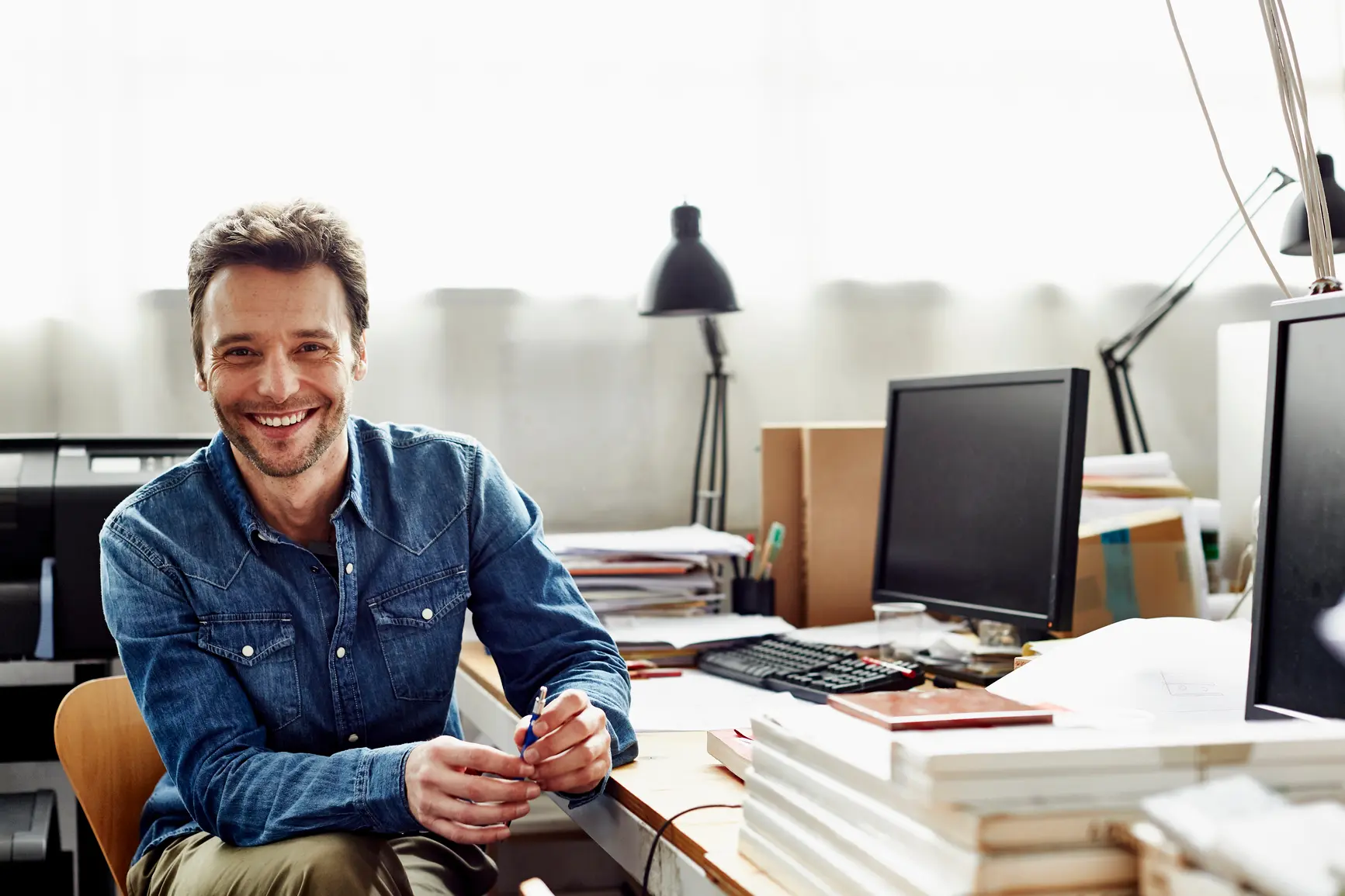 A man smiles into the camera. To his right you can see a full desk with a stack of papers and a PC.