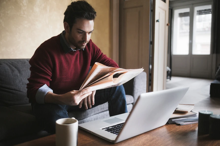 A man is sitting on a couch with a newspaper in his hand. On the table in front of him are a laptop and a coffee mug.