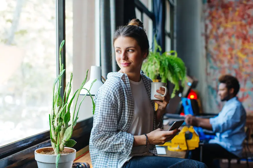 A woman sits on a windowsill, holding a cup in one hand and a smartphone in the other, looking out the window.