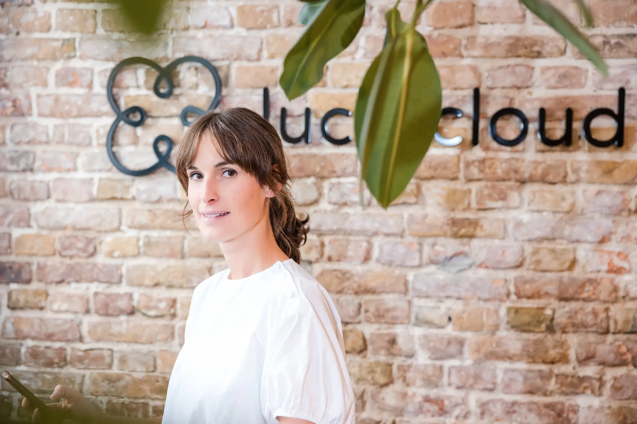 A woman smiles into the camera, the logo of luckycloud can be seen on the stone wall behind her.