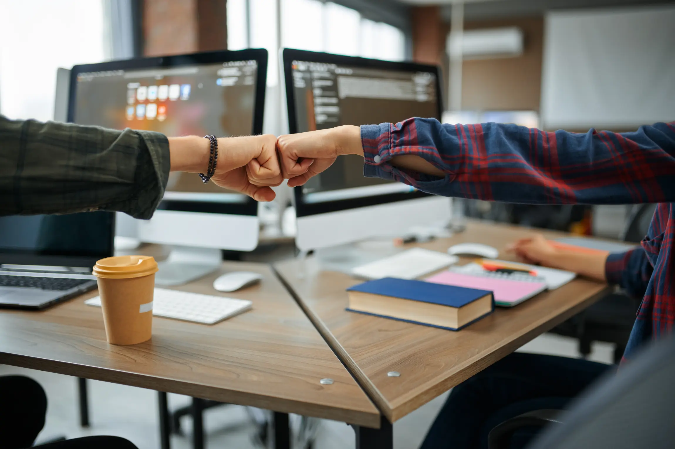 Two people give each other a fist bump at their shared desk. Their PCs can be seen in the background.