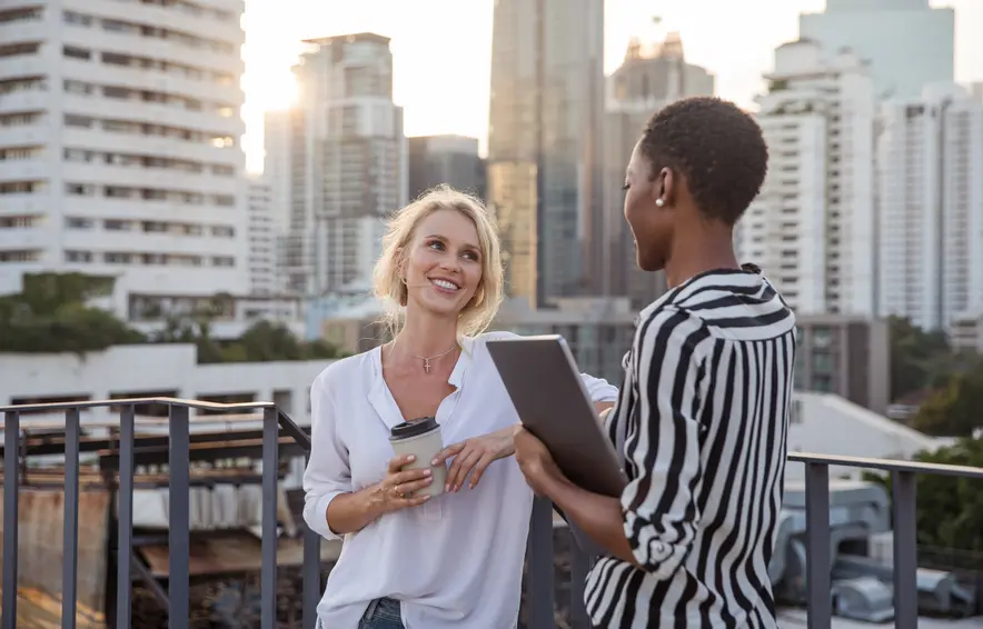 Two happy people are standing on a roof terrace with coffee and laptop in hand. In the background you can see skyscrapers.