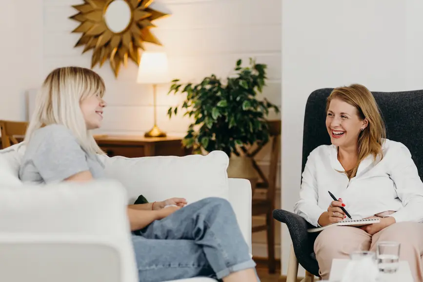 Two women are each sitting in an armchair and laughing at each other. One woman has a notebook on her lap.