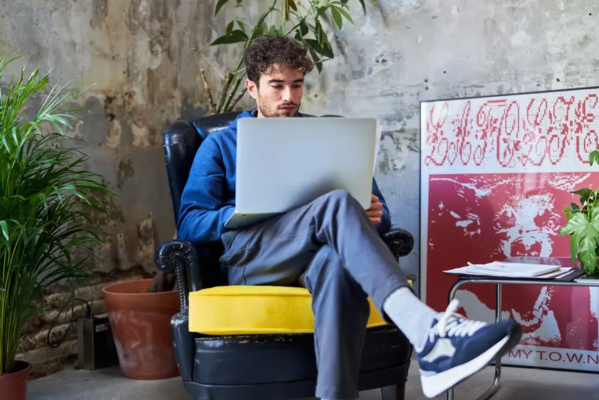 Ein junger Mann sitzt in einem Sessel. Auf seinem Schoss steht ein Laptop.