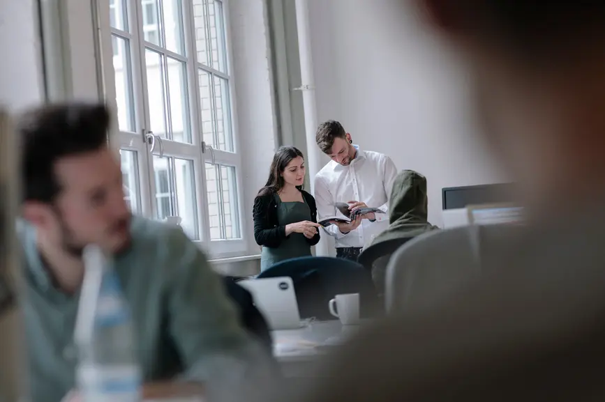 Im Hintergrund stehen zwei Personen, die durch eine Broschuere blaettern. Im Vordergrund sind zwei Kollegen verschwommen zu sehen.