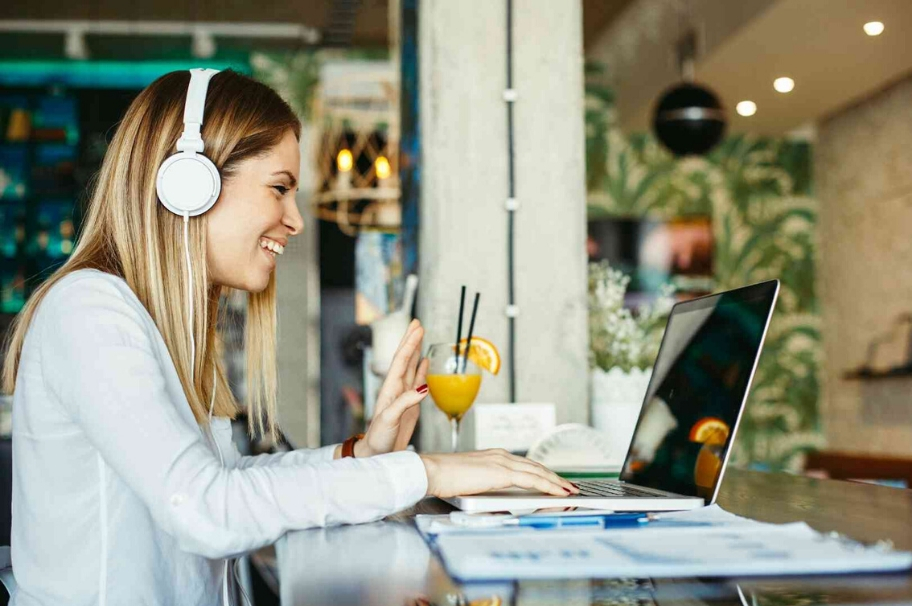 A woman sits with headphones in front of her laptop and waves into the screen. Next to her is a cocktail glass