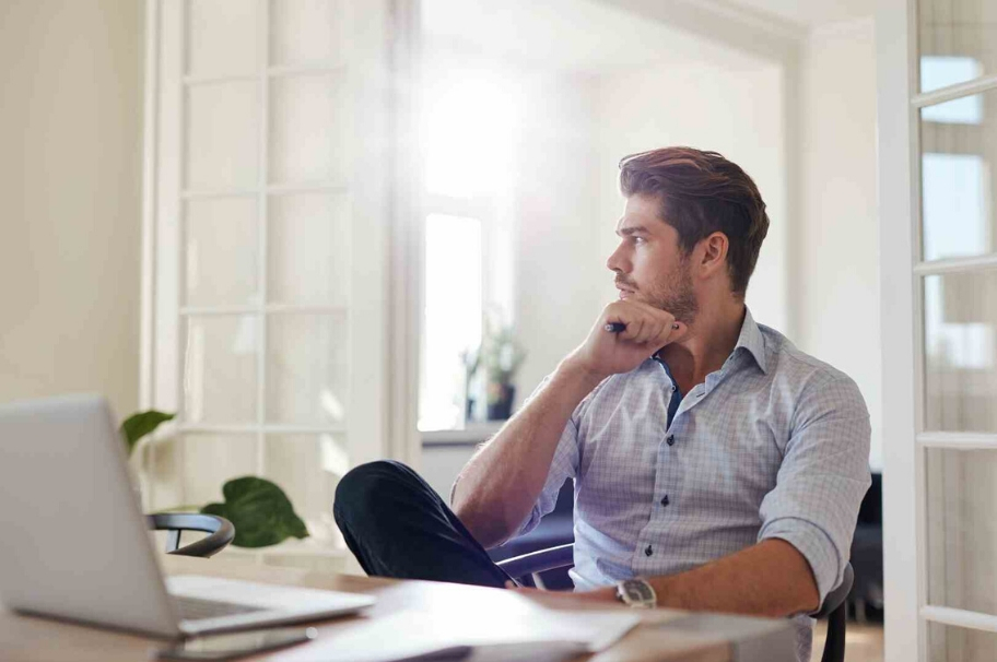 A man sits in front of his laptop and look thoughtfully towards the window