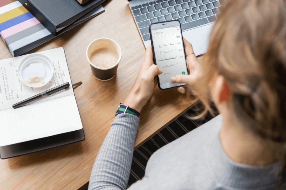 Bird's eye view of a desk with laptop, coffee mug and notepad. In her hand the woman is holding a smartphone.