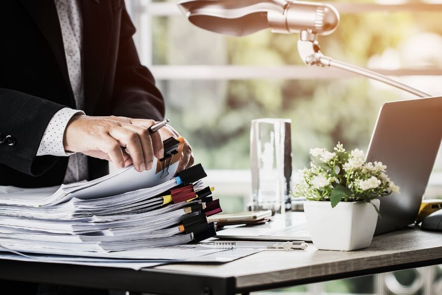 Close-up of a table with a stack of papers on it. A person in the background is working on this