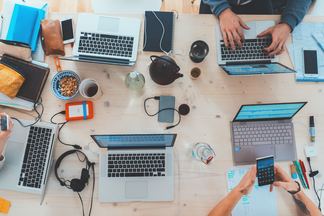 Vogelperspektive auf einen großen Schreibtisch mit mehreren Laptops, Handys, Ladegeraeten und Co.