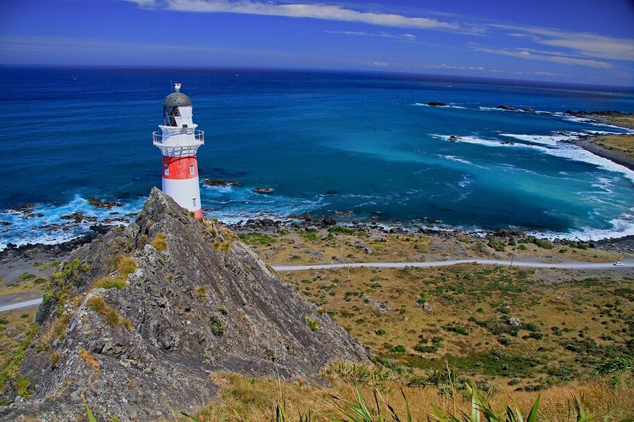 A red and white lighthouse stands in front of the coast of an azure sea.