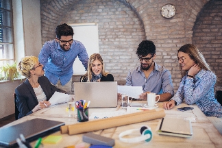 A team sits and stands around a desk and looks together at laptop and notes.