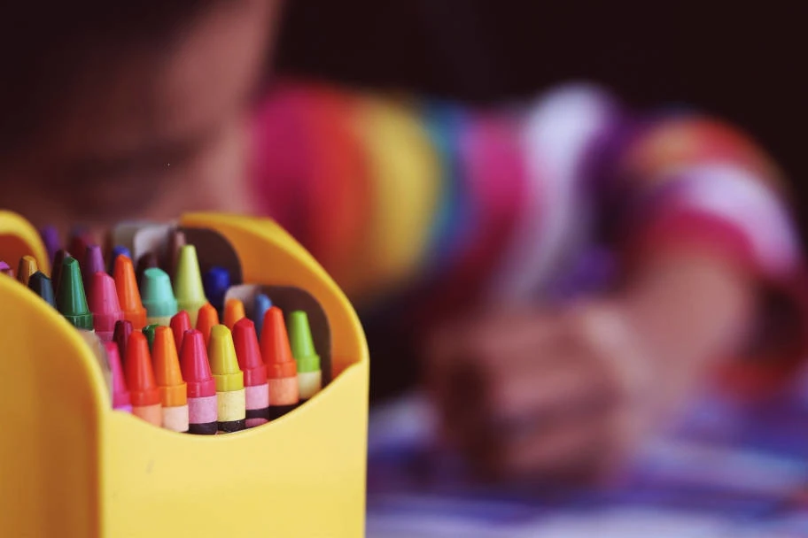 Im Vordergrund des Bildes ist ein Stiftehalter mit Buntstiften zu sehen. Der verwaschene Hintergrund zeigt ein Kind.