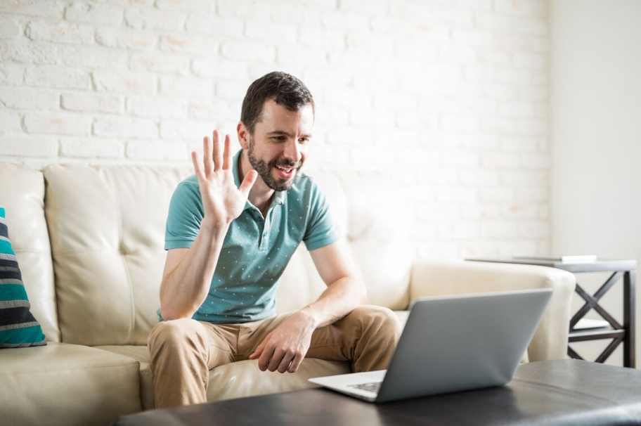 A man is sitting on a sofa, his laptop in front of him. Into which he waves